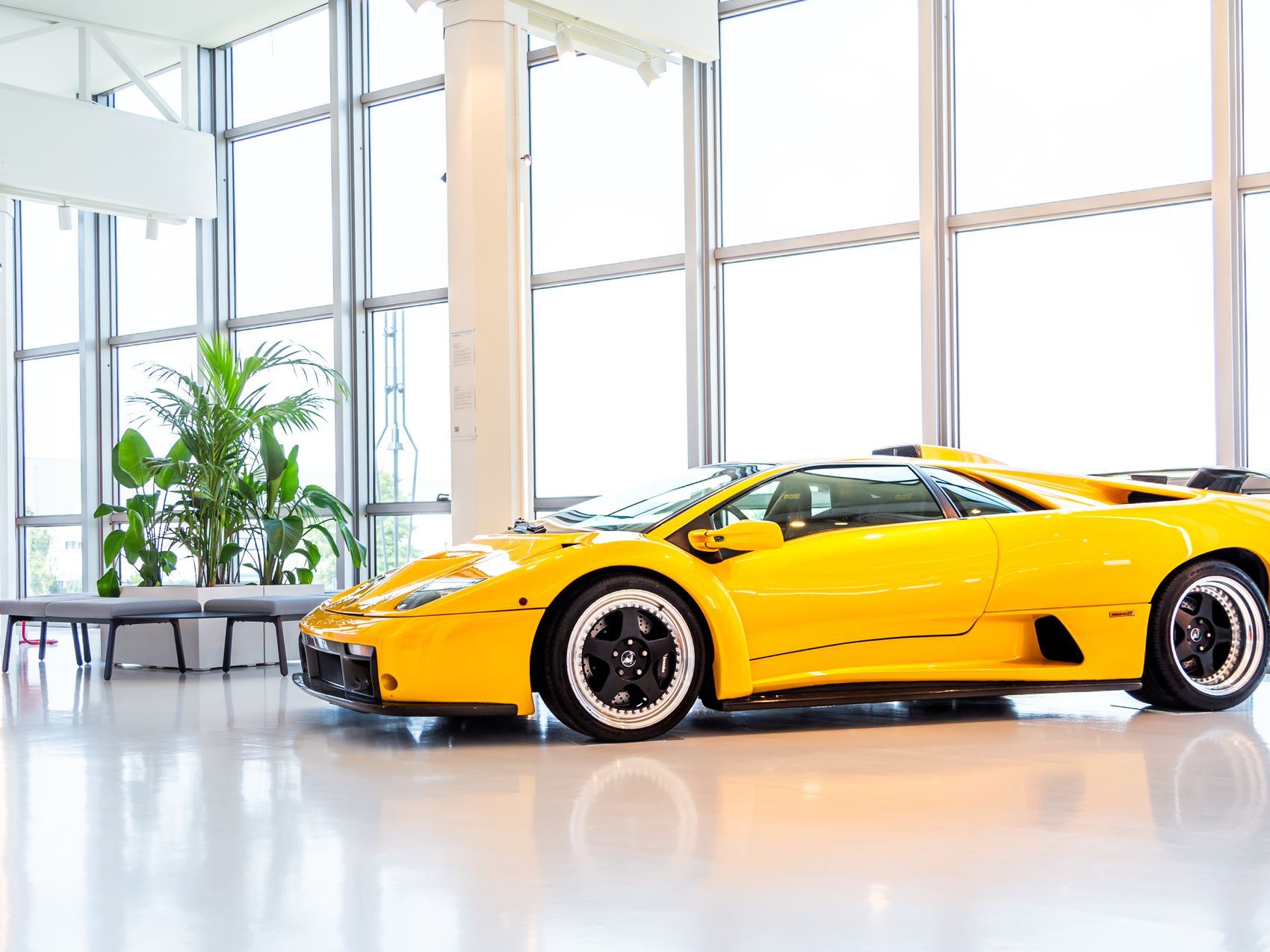 A yellow sports car on a glossy light floor in a bright glazed showroom.